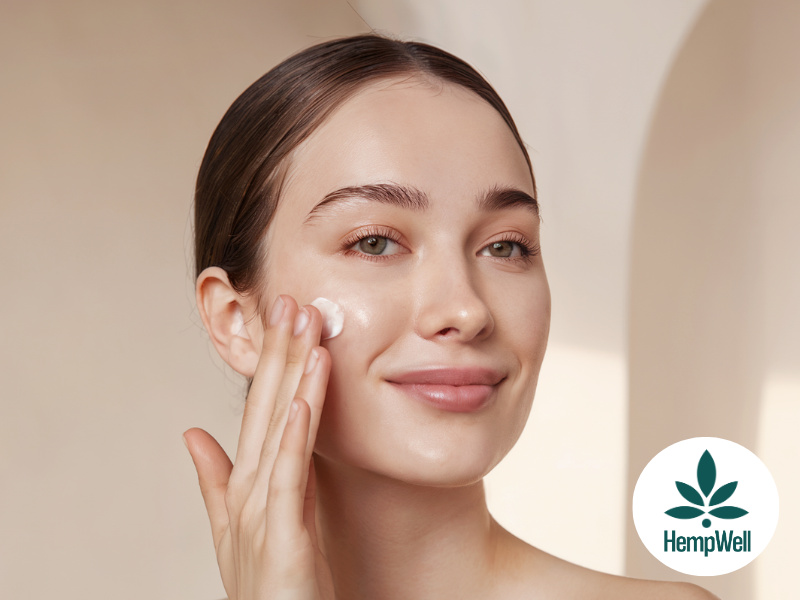 Woman applying moisturizer to her cheek with a cotton pad; HempWell logo visible in the bottom-right corner.