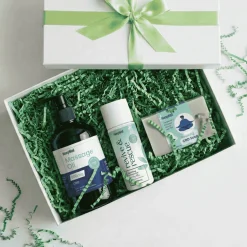 Gift box with green ribbon containing CBD wellness items: massage oil, revive & rescue spray, and CBD soap on green shredded filler.