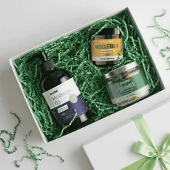 CBD wellness gift set with massage oil, muscle balm, and gummies in a white gift box with green filler material.
